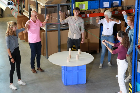 Eight people work together to stack six miniature blocks into a single, stable tower.