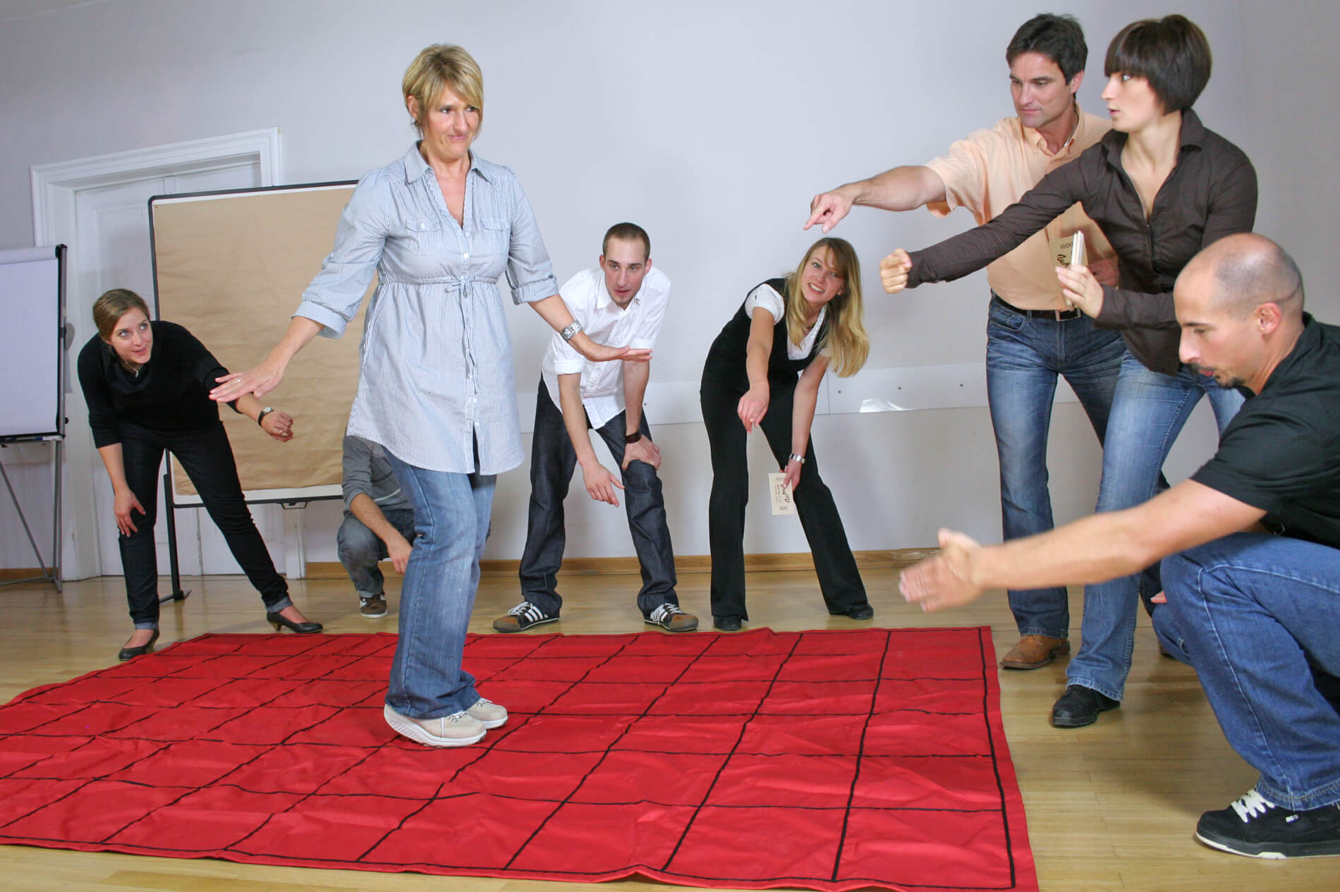 A work team tells a woman how to walk across a red floor mat in an activity called The Maze.