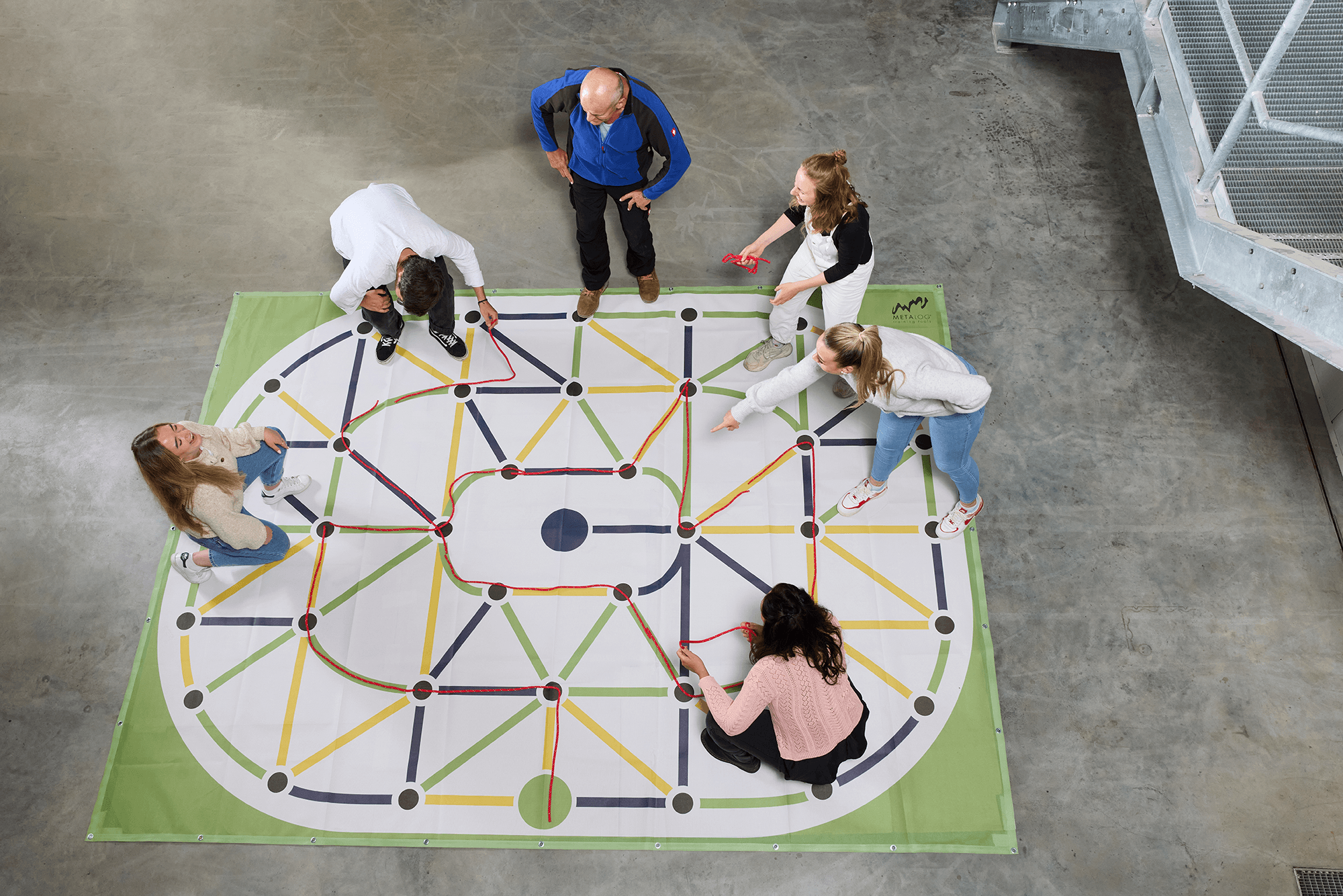Six adults are laying red strings on a color maze set on a concrete floor 