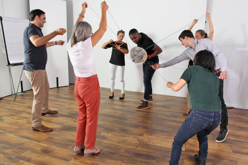 Seven adults surround PerspActive, each holding strings to move the sphere, a team building tool 