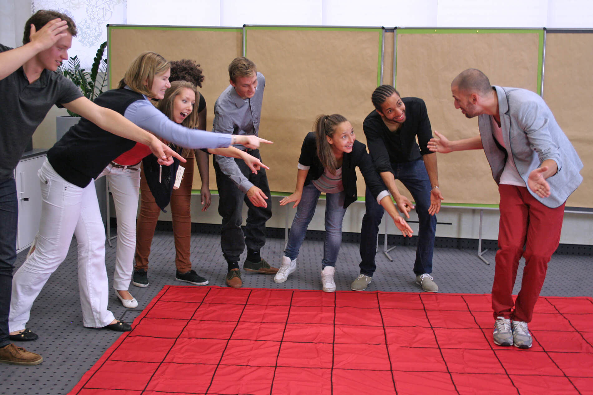 A man standing on a red floor mat called The Maze receives movement recommendations from coworkers.